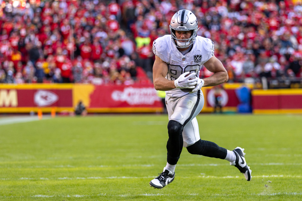 Brock Bowers, in white uniform with a silver helmet, runs upfield with the football tucked in front of him.